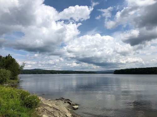 A serene lake scene with calm water, lush greenery, and a sky filled with fluffy clouds.