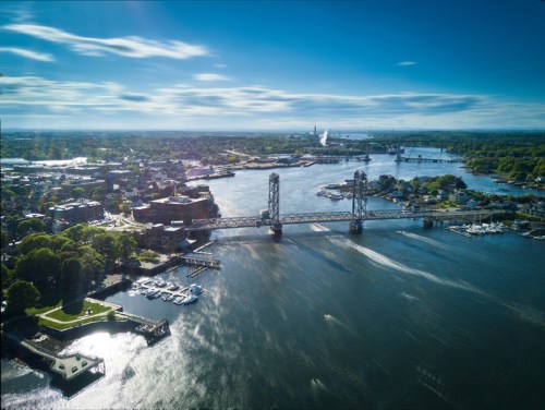 Aerial view of a river with a bridge, surrounded by greenery and buildings under a clear blue sky.