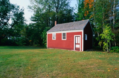 A small red house with white trim sits in a grassy area surrounded by trees in autumn foliage.