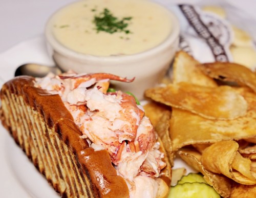 A lobster roll with creamy filling, served with a bowl of chowder, potato chips, and pickles on the side.