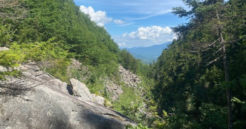 A scenic view of a rocky valley surrounded by lush green trees and mountains under a blue sky with fluffy clouds.