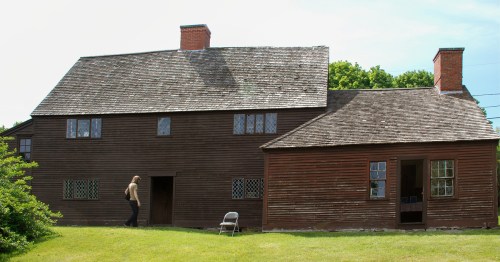 A person walks past a large, rustic wooden house with a sloped roof and multiple windows on a sunny day.