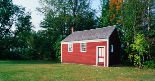 A small red wooden house surrounded by trees and grass, with a white door and windows.