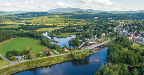 Aerial view of a scenic landscape featuring a river, green fields, and a small town surrounded by mountains.