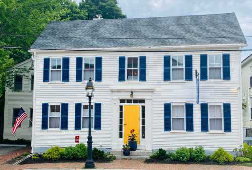 A charming white house with blue shutters, a yellow door, and an American flag, surrounded by greenery and brick pathway.