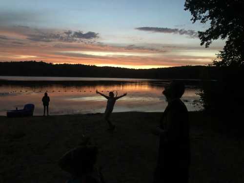 Silhouettes of people on a beach at sunset, with a calm lake and colorful sky in the background.