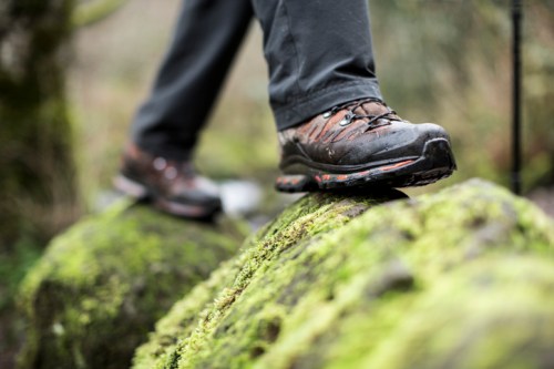 A close-up of a hiker's boot stepping on a moss-covered rock in a natural setting.