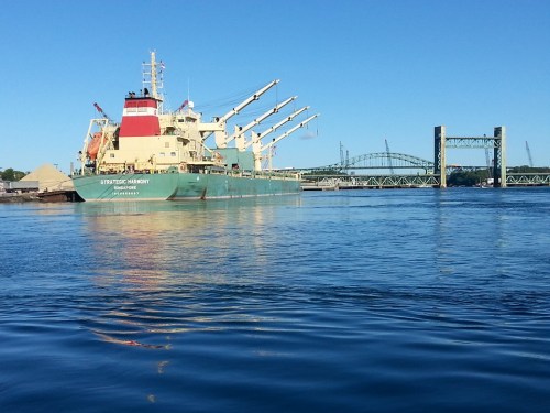 A large cargo ship docked near a bridge, with clear blue skies and calm water reflecting the scene.