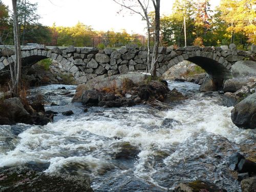 A stone bridge arches over a rushing river, surrounded by trees and rocky terrain in a serene natural setting.