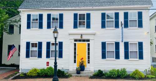 A charming white house with blue shutters and a bright yellow door, featuring a lamp post and an American flag.