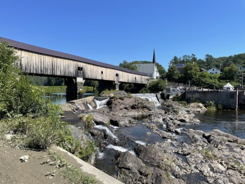 A wooden covered bridge spans a river with small waterfalls and rocky banks under a clear blue sky.
