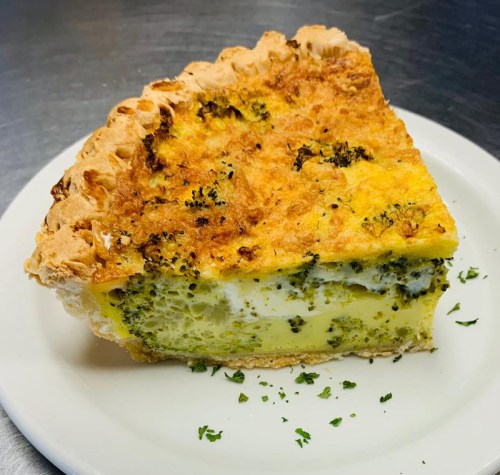 A slice of broccoli quiche with a golden crust, served on a white plate, garnished with parsley.