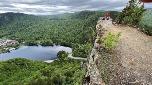 A person sits on a rocky ledge overlooking a lush green valley and a winding lake below, under a cloudy sky.
