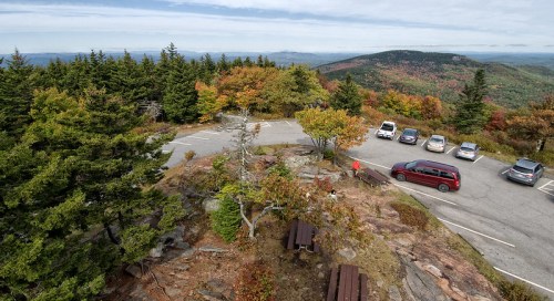 A scenic overlook with autumn foliage, parking area, and distant mountains under a cloudy sky.