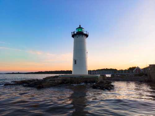 A tall white lighthouse with a green light stands by the water at sunset, surrounded by rocky shore and calm waves.