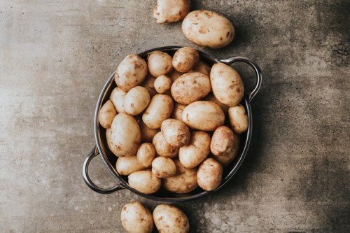 A metal bowl filled with various sizes of fresh, unpeeled potatoes on a textured surface.