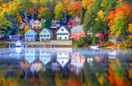 Colorful houses reflect in a calm lake surrounded by vibrant autumn foliage. Mist hovers over the water.