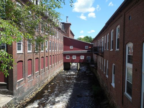 Historic red brick buildings alongside a flowing river under a blue sky with scattered clouds.