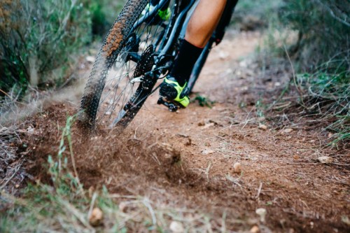 A close-up of a mountain bike tire kicking up dirt on a trail, with a rider's foot visible in motion.