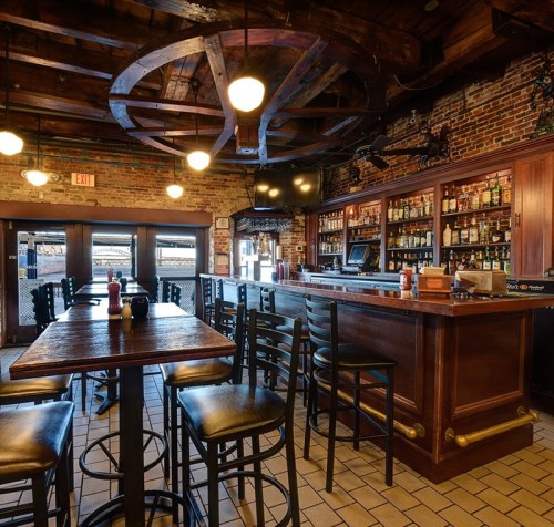 Cozy bar interior with wooden beams, a brick wall, and a well-stocked counter, featuring tables and chairs.