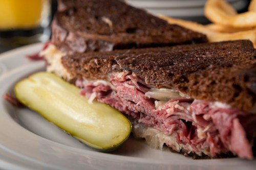 Close-up of a sliced sandwich with layers of meat and sauerkraut, served with a pickle on the side.