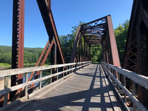 A rustic metal bridge with wooden planks, surrounded by greenery and blue skies, casting shadows on the ground.