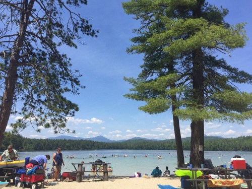 A sunny beach scene with people enjoying the water, surrounded by trees and mountains in the background.