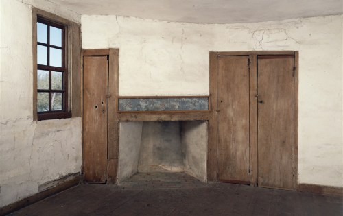 Interior of a rustic room featuring a simple fireplace, wooden doors, and a window with natural light.