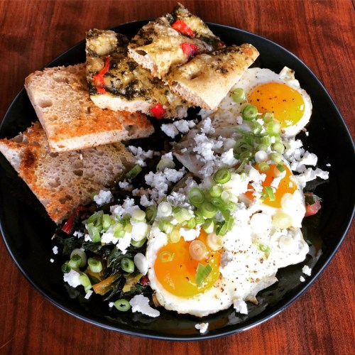 A black plate with two sunny-side-up eggs, crumbled feta, green onions, and slices of toasted bread.
