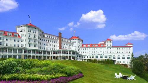 A grand white hotel with red roofs, set on a lush green lawn under a blue sky with fluffy clouds.
