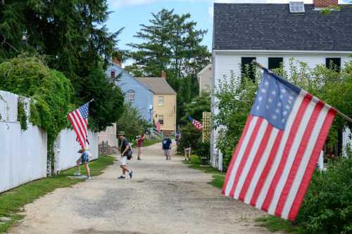 A quaint street lined with houses and American flags, with people walking along a gravel path.