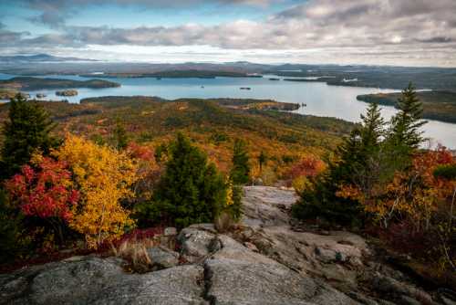 A scenic view of a lake surrounded by vibrant autumn foliage and distant mountains under a cloudy sky.