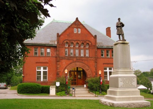 Historic red brick library building with a statue of a soldier in front, surrounded by greenery and flags.
