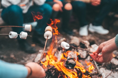 A close-up of hands roasting marshmallows over a campfire, with people sitting around in the background.
