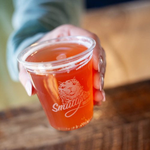 A hand holding a clear cup of amber beer with a logo that reads "Smuttynose Brewing."