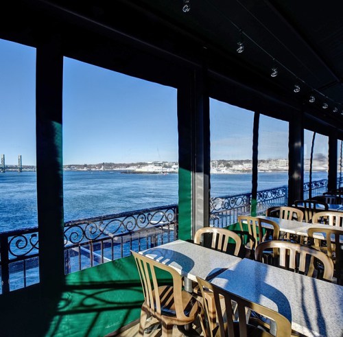 View from a restaurant with tables overlooking a calm waterfront and distant shoreline under a clear blue sky.