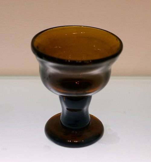 A small, elegant glass cup with a dark amber color and a sturdy base, set against a light background.