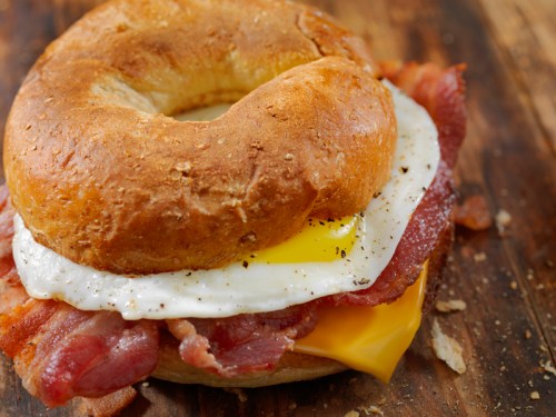 A toasted bagel sandwich with bacon, a fried egg, and melted cheese on a wooden surface.