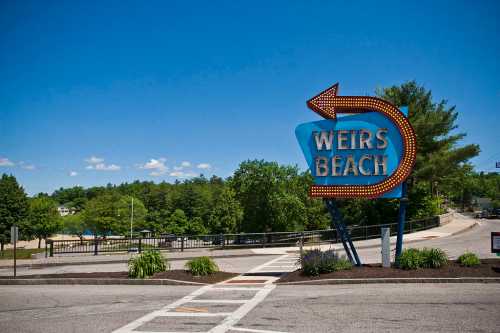 Sign for Weirs Beach with a blue arrow, surrounded by greenery and a clear blue sky.