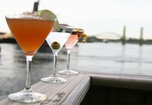 Three cocktails on a wooden railing, with a scenic waterfront and bridge in the background.