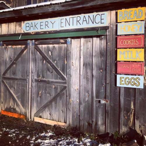 A rustic bakery entrance with wooden doors and colorful signs for bread, honey, cookies, milk, maple syrup, and eggs.