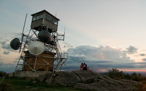 A lookout tower with large satellite dishes, overlooking a scenic landscape at sunset, with two people sitting nearby.