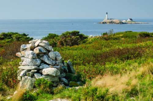 A stone cairn in the foreground with a lighthouse on a rocky island and calm waters in the background.