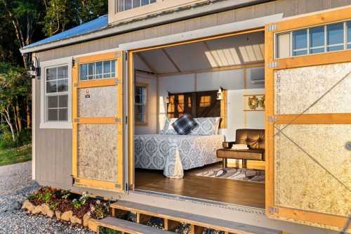 Cozy bedroom with a bed and sofa, open doors revealing a serene outdoor view in a tiny house setting.