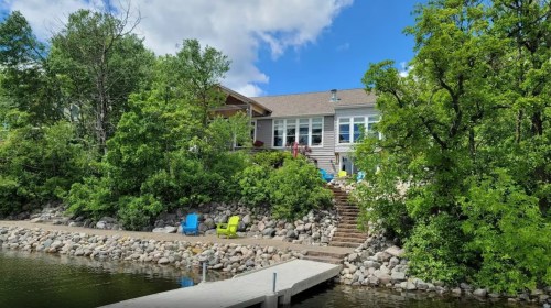 A lakeside house surrounded by greenery, with a dock and colorful chairs along the shore under a blue sky.