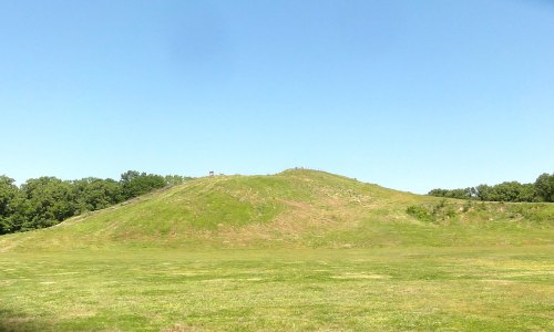 A grassy hill rises against a clear blue sky, surrounded by a green field and trees in the background.