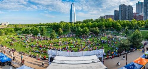 A vibrant outdoor event with a large crowd on green grass, featuring tents and the Gateway Arch in the background.