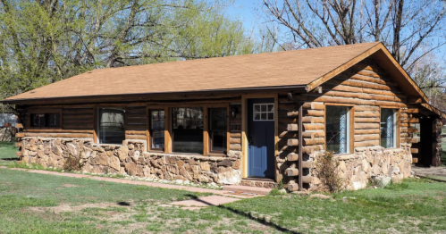 A rustic log cabin with a stone foundation, large windows, and a blue front door, surrounded by green grass and trees.