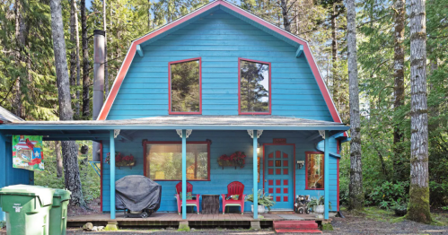 A vibrant blue cabin surrounded by trees, featuring a porch with red chairs and green trash bins in front.