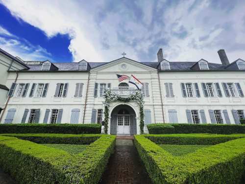 A historic building with a white facade, surrounded by neatly trimmed hedges and flags, under a cloudy sky.